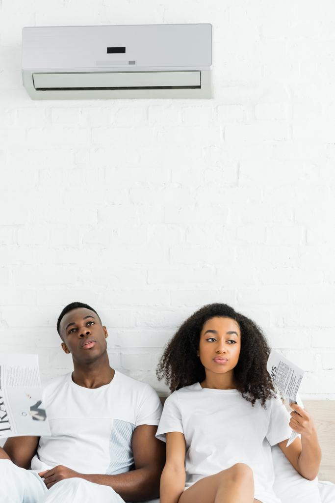 fatigué afro-américain homme et femme avec des journaux dans les mains assis sur le lit dans la chambre
 - Photo, image