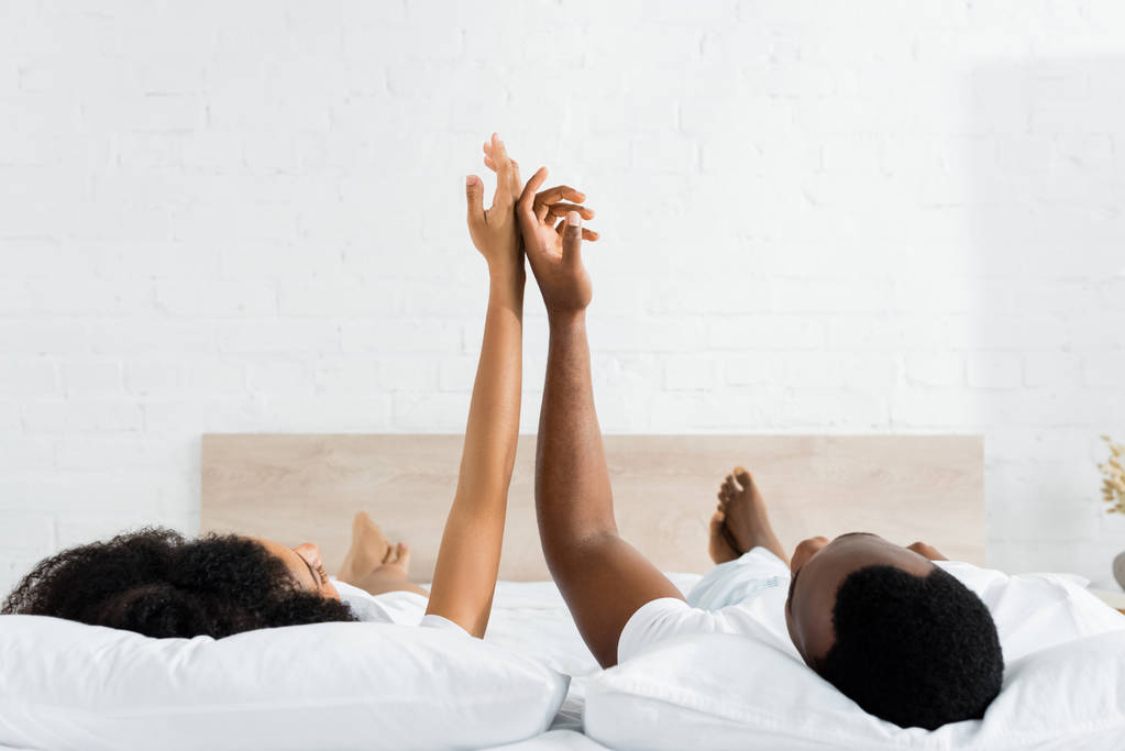 счастливый африканский американец, касающийся рук с девушкой, лежащий на белой кровати в спальне
 - Фото, изображение