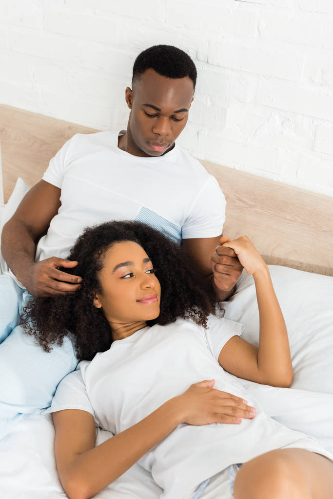 Widok z dużym kątem African American mężczyzna trzyma ręce z piękną dziewczyną, leżąc na łóżku - Zdjęcie, obraz