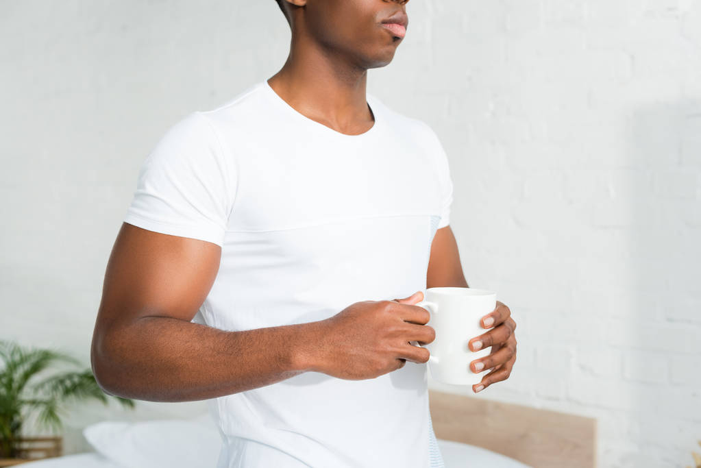 bijgesneden beeld van African American man Holding Cup in handen, staande in witte kamer - Foto, afbeelding