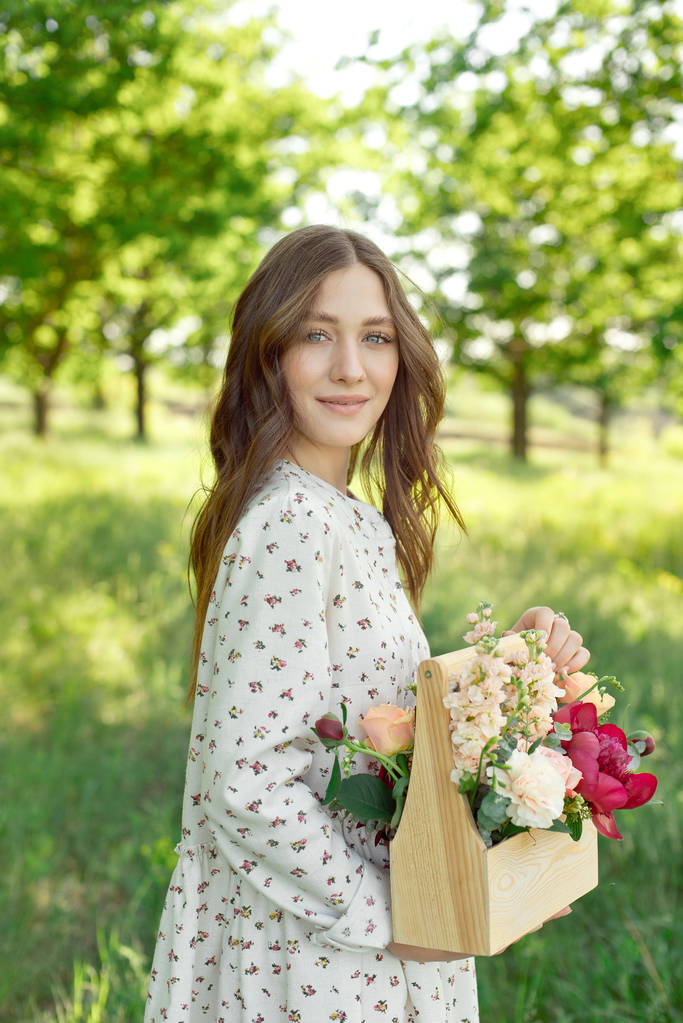 Mitad retrato de una encantadora mujer positiva vestida con largos vestidos blancos de verano con una sonrisa feliz en el fondo de un prado verde en las manos de una cesta de madera con peonías, rosas, lirios verdes
 - Foto, Imagen