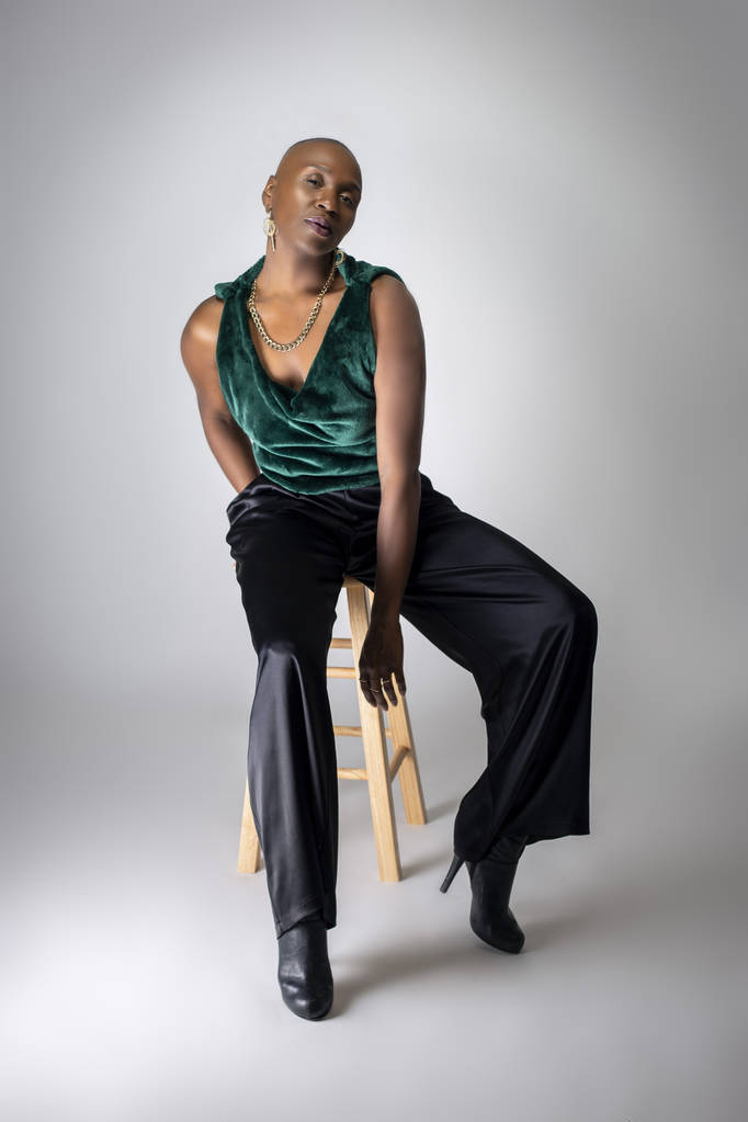Modèle de mode féminine afro-américaine noire portant une tenue verte tendance et des talons assis sur un tabouret. Elle exhibe avec confiance une coiffure chauve audacieuse
.   - Photo, image