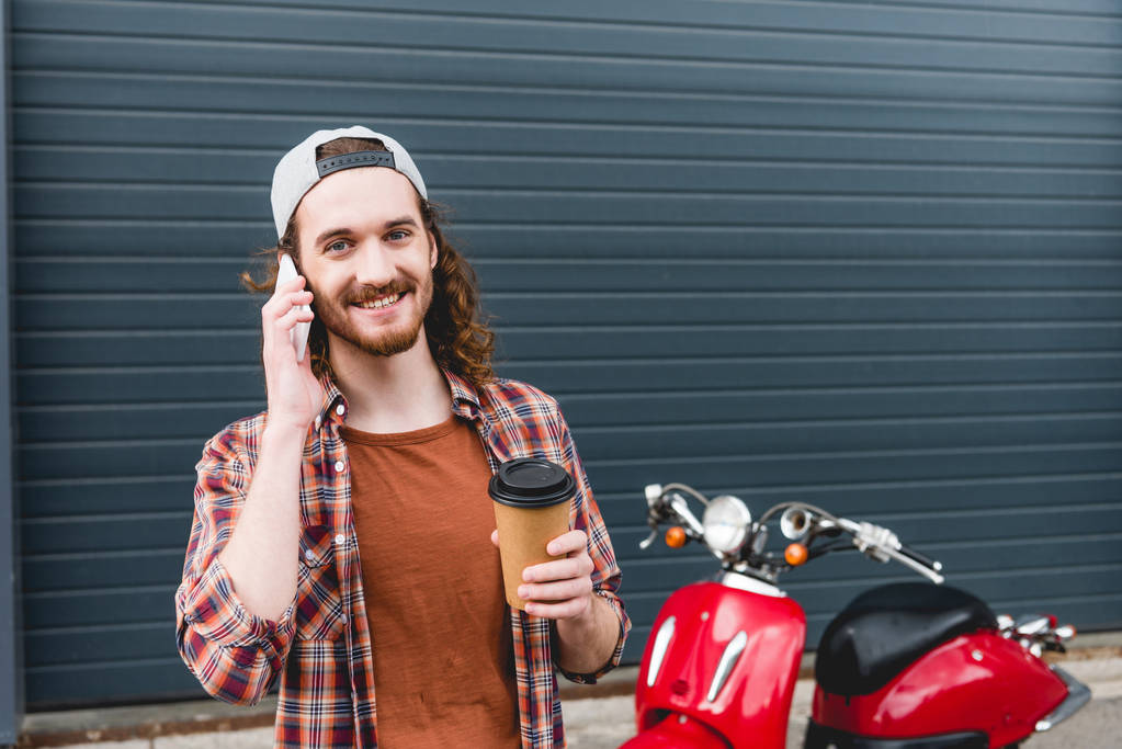 mladý muž mluví na smartphone, drží papírový kelímek s kávou a stojí blízko červené koloběžky  - Fotografie, Obrázek