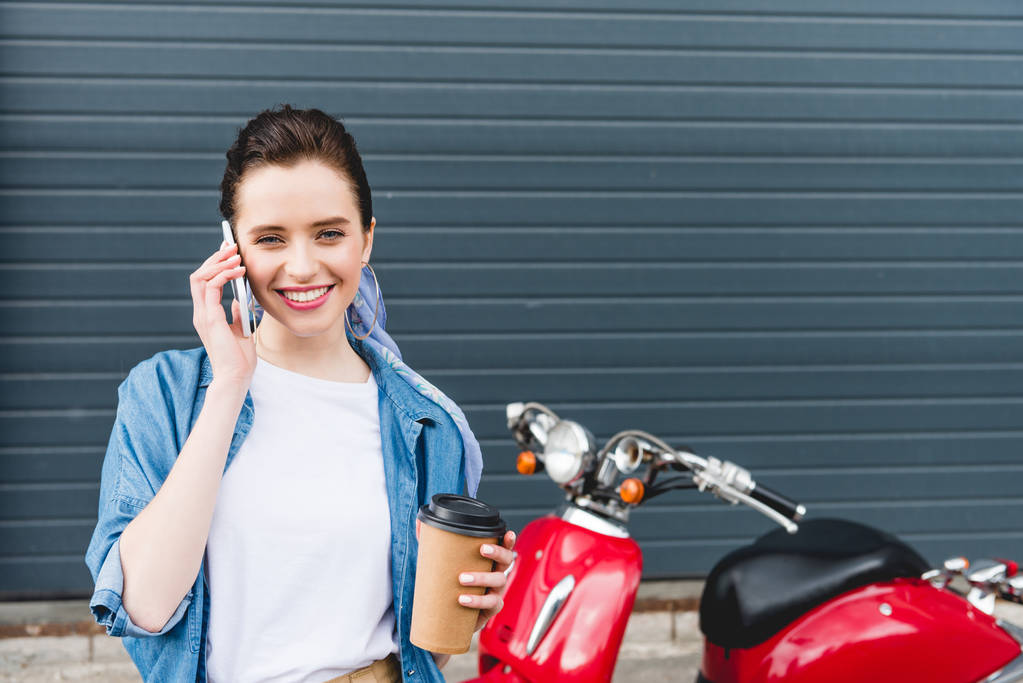 Vorderansicht eines schönen Mädchens, das in der Nähe eines roten Motorrollers steht, Pappbecher mit Kaffee in der Hand hält und auf dem Smartphone spricht - Foto, Bild