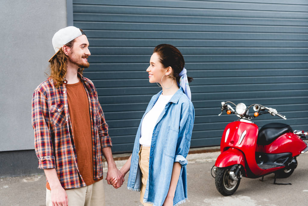 jonge man en meisje kijken elkaar aan en hand in hand met rode scooter op de achtergrond - Foto, afbeelding