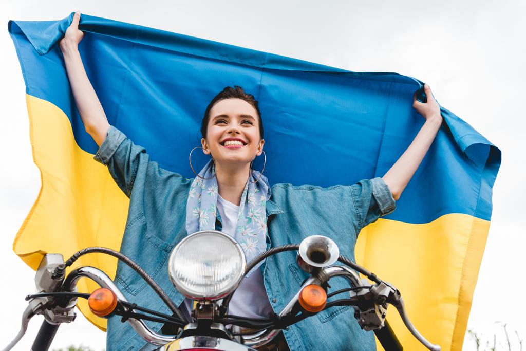 vista de ángulo bajo de hermosa chica sentada en scooter, sonriendo y sosteniendo la bandera de Ucrania
 - Foto, Imagen