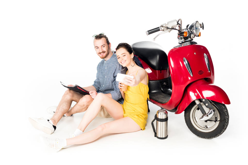 joven hombre y chica sentado en el suelo cerca de scooter rojo y tomar selfie aislado en blanco
 - Foto, imagen