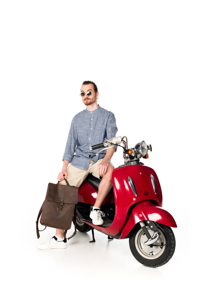 kırmızı scooter üzerinde oturan ve beyaz izole sırt çantası tutan yakışıklı şık genç adam tam uzunlukta görünümü - Fotoğraf, Görsel