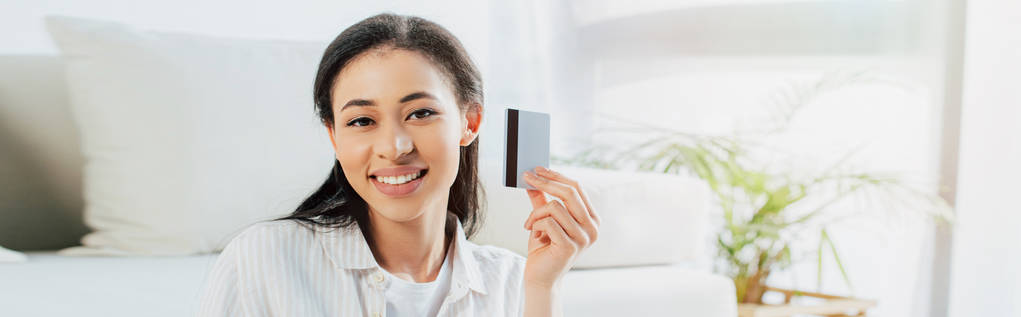 tiro panorâmico de mulher latina alegre segurando cartão de crédito e olhando para a câmera
 - Foto, Imagem