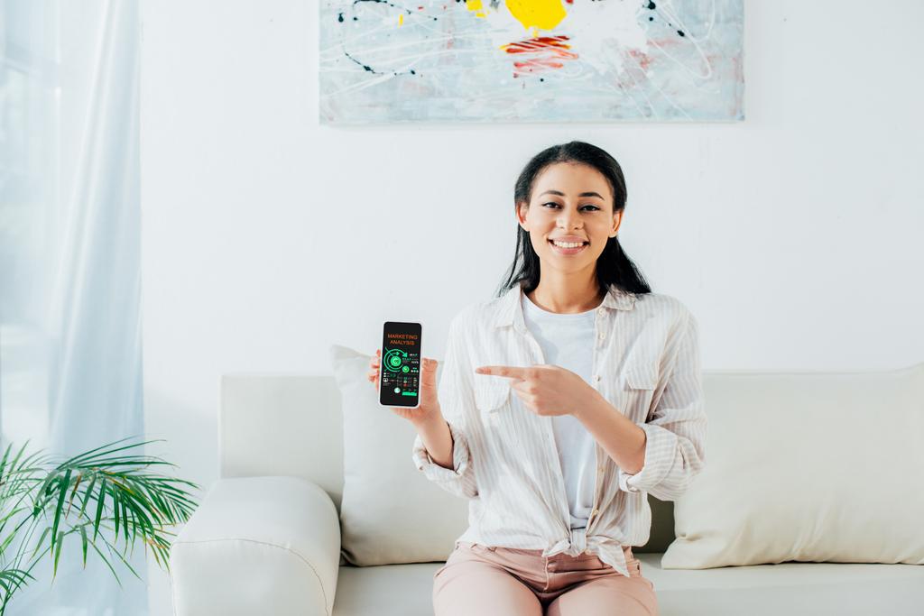 mujer latina bonita apuntando con el dedo al teléfono inteligente con aplicación de análisis de marketing en la pantalla
 - Foto, imagen