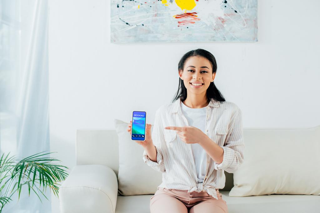 mujer latina alegre apuntando con el dedo al teléfono inteligente con aplicación de reserva en la pantalla
 - Foto, Imagen