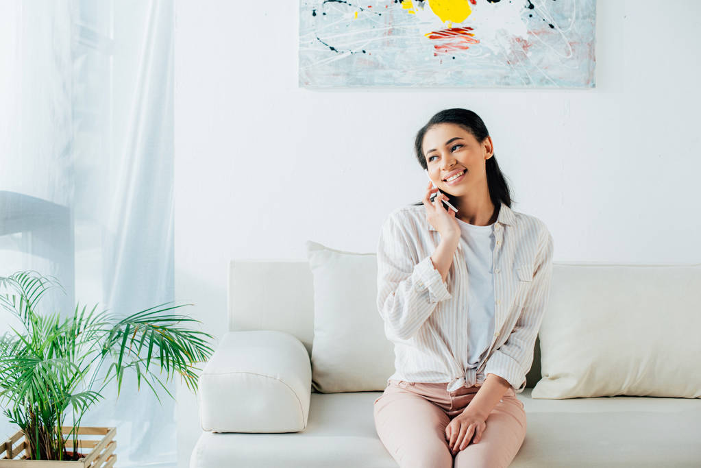 hermosa mujer latina hablando en el teléfono inteligente mientras está sentada en el sofá y mirando hacia otro lado
 - Foto, imagen