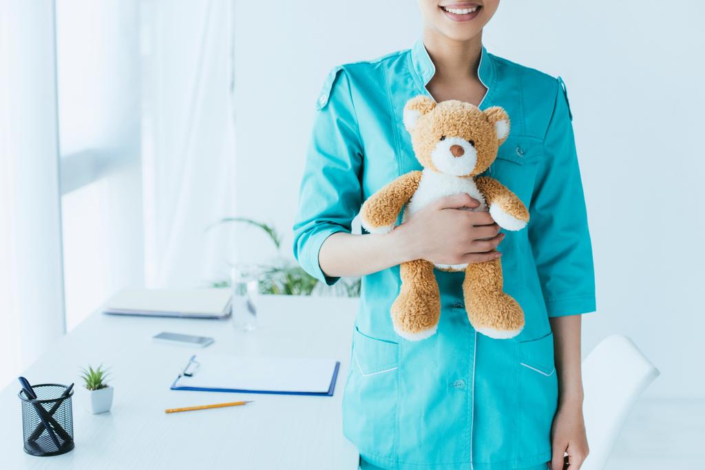 частковий вид на молодого латинського лікаря, який тримає плюшевого ведмедя, стоячи біля робочого місця
 - Фото, зображення