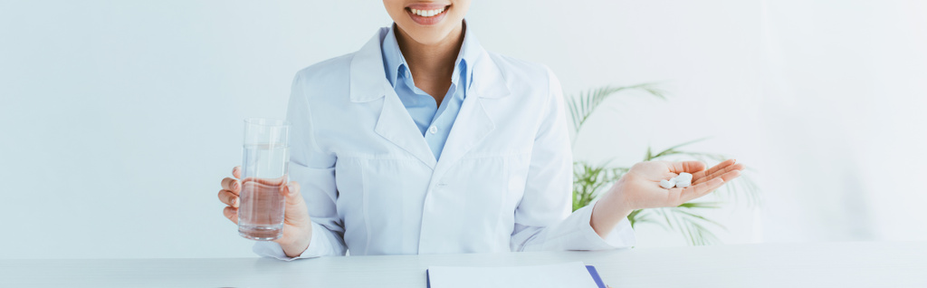 πανοραμική φωτογραφία χαμογελαστή γιατρού με χούφτα χάπια και ποτήρι νερό - Φωτογραφία, εικόνα