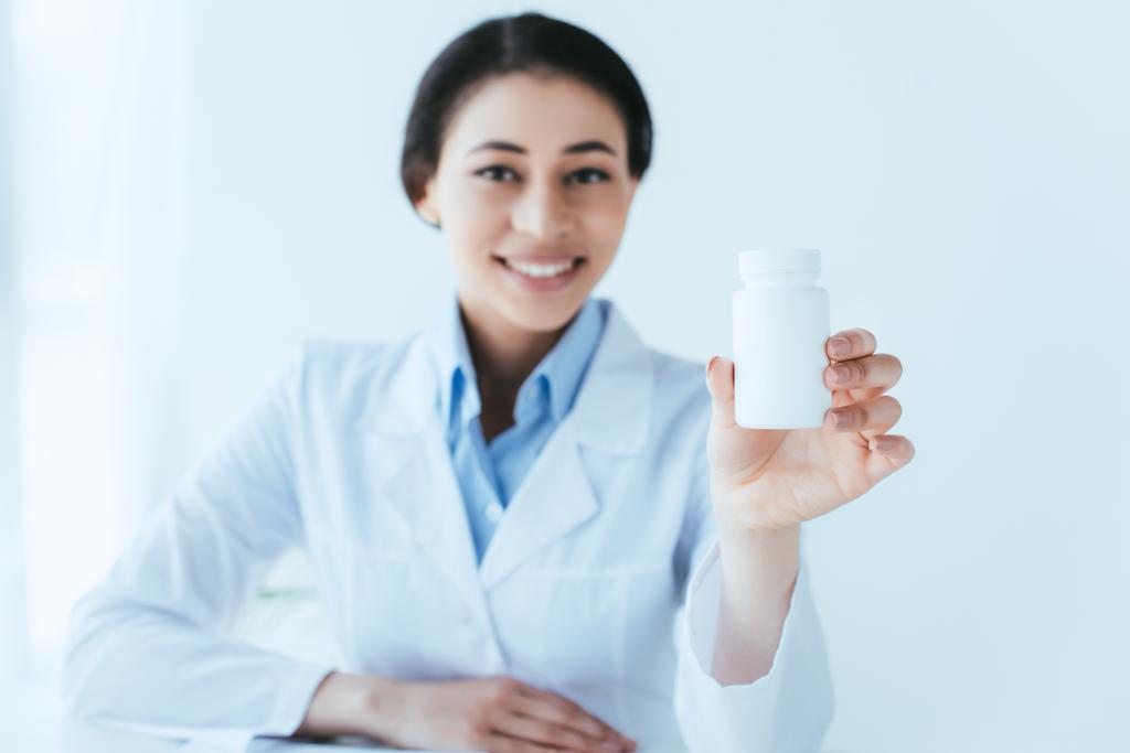selective focus of cheerful latin doctor holding pills container and smiling at camera - Photo, Image