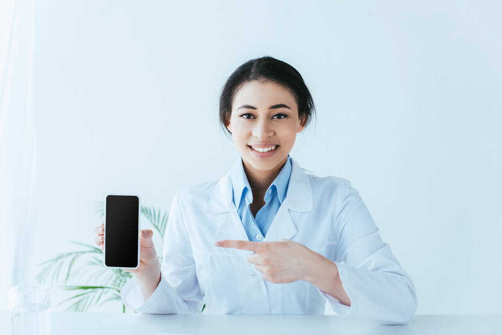 привлекательный латинский врач улыбается в камеру и указывает пальцем на смартфон с пустым экраном
 - Фото, изображение