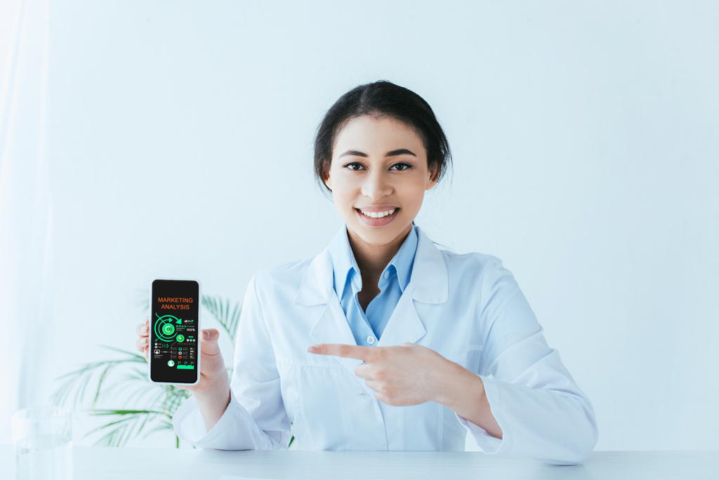 młody latynoski lekarz wskazujący palcem na smartfona z analizą marketingową na ekranie - Zdjęcie, obraz