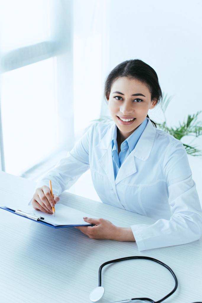 attractive latin doctor writing on clipboard while smiling at camera in office - Photo, Image