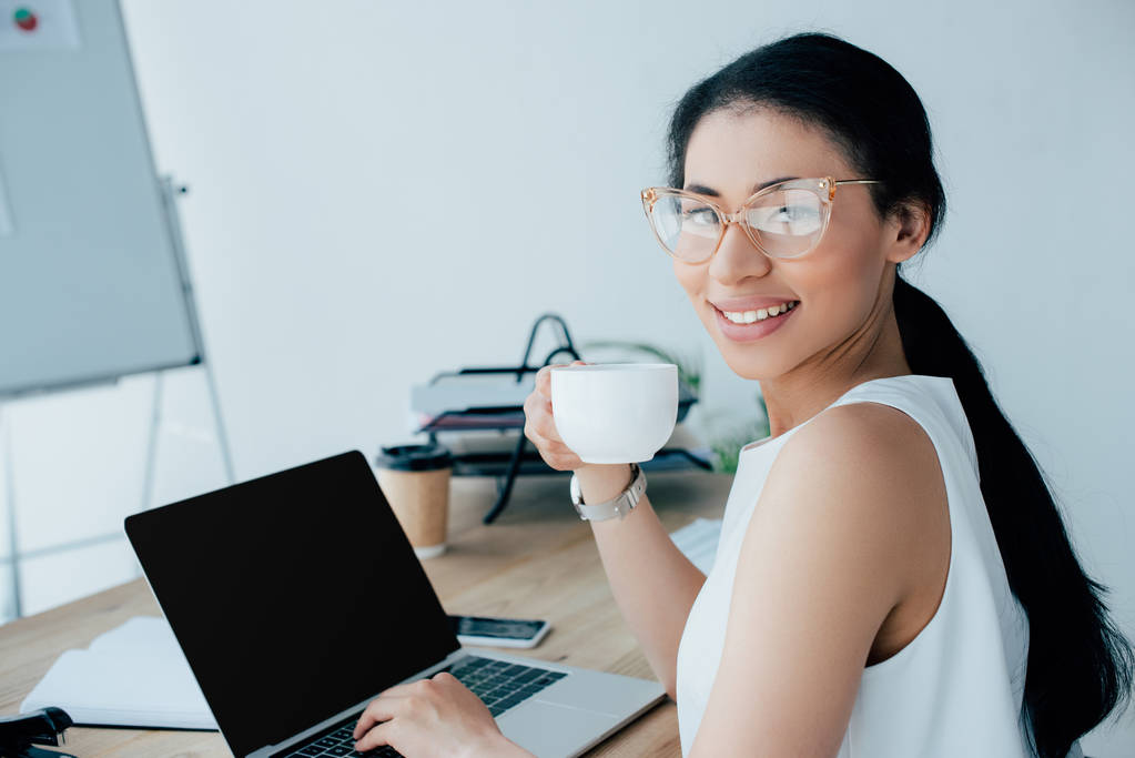 mulher de negócios latina alegre segurando xícara de café e usando laptop enquanto sorri para a câmera
 - Foto, Imagem