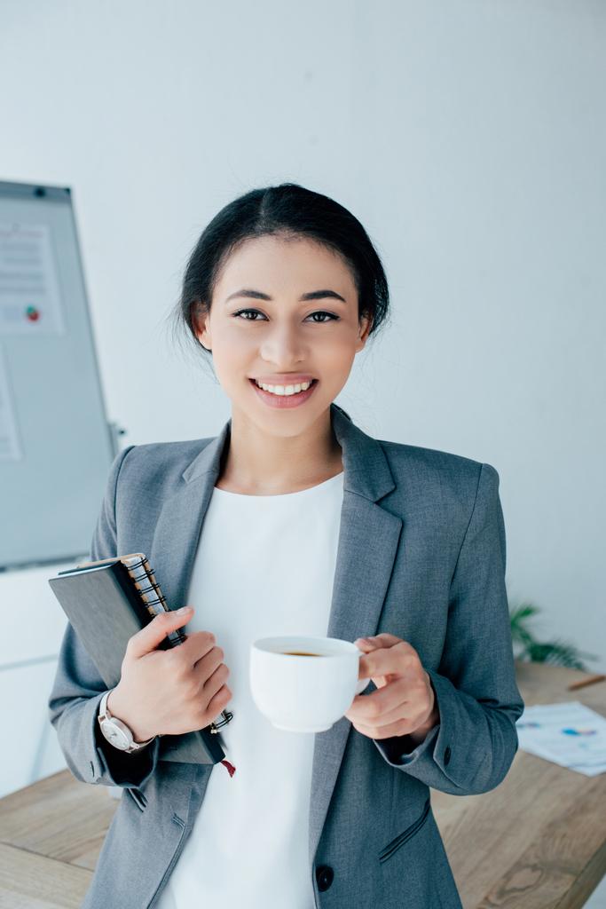 Ładna Łacińska businesswoman trzymając Notebooki i filiżankę kawy podczas uśmiechu w aparacie - Zdjęcie, obraz