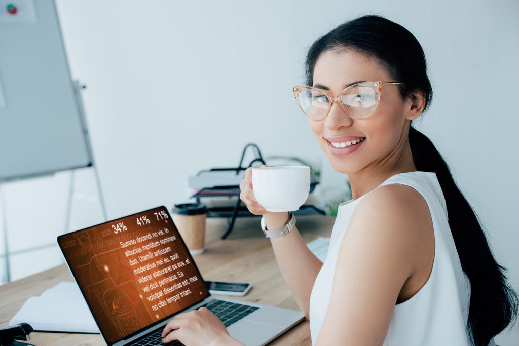 χαμογελαστή Λατινική επιχειρηματίας κρατώντας κούπα καφέ, ενώ χρησιμοποιεί φορητό υπολογιστή με πληροφορίες κειμένου στην οθόνη - Φωτογραφία, εικόνα