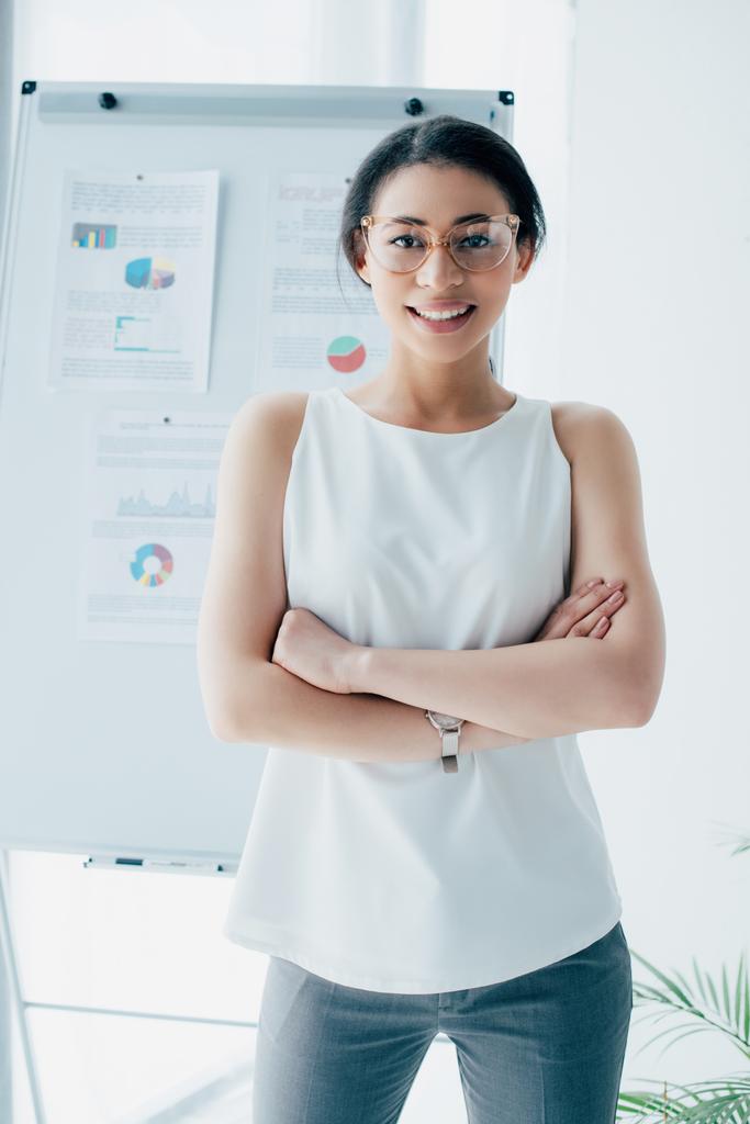 Ładna Łacińska businesswoman w okularach uśmiechniętych w aparacie stojąc z skrzyżowanymi ramionami w biurze - Zdjęcie, obraz