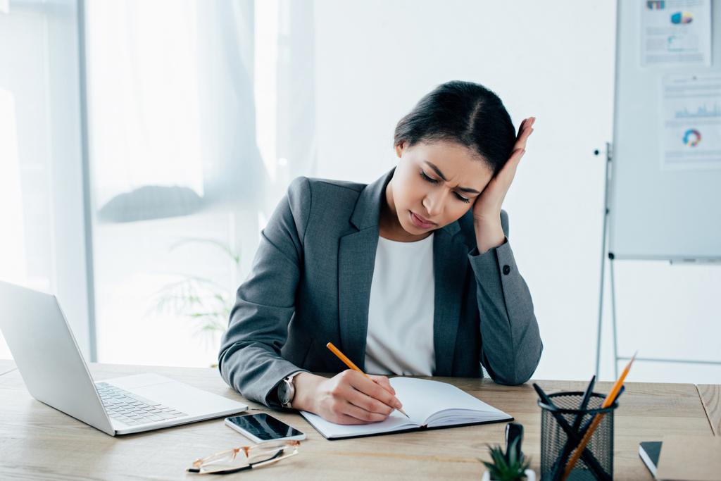 wyczerpane Latin businesswoman piśmie w Notatniku siedząc na biurku i trzymając rękę w pobliżu głowy - Zdjęcie, obraz