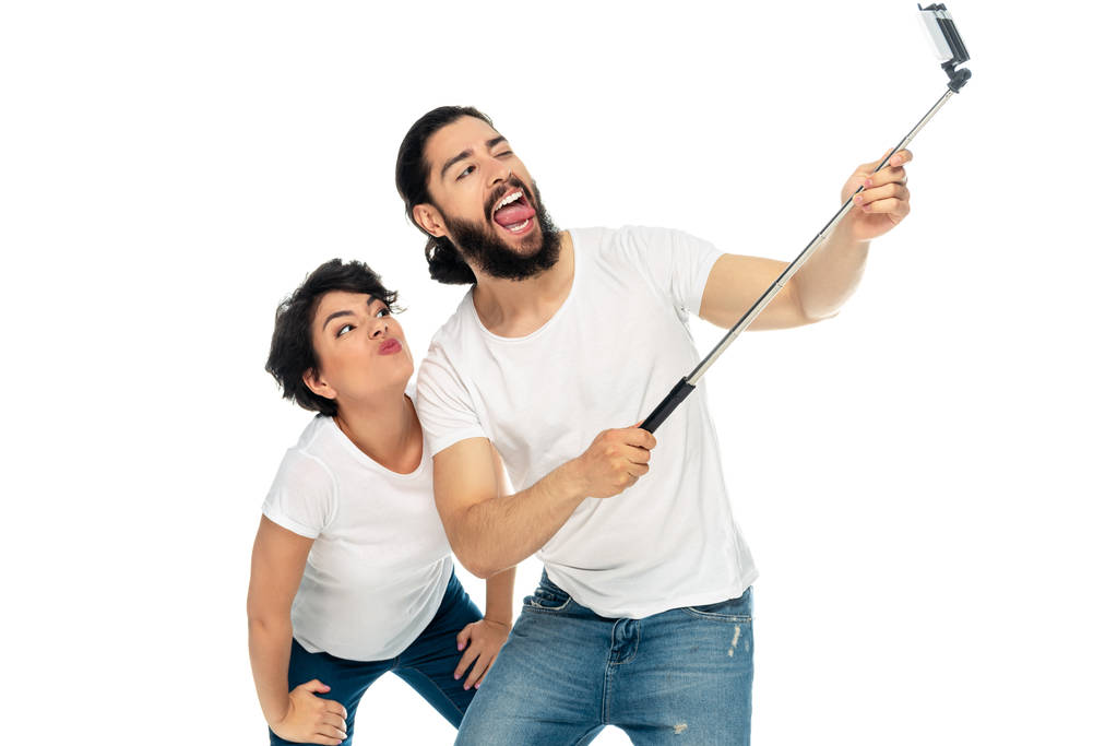 szczęśliwy łaciński człowiek pokazując język w pobliżu kobiety brunetka i trzymając Selfie Stick podczas robienia selfie na białym tle  - Zdjęcie, obraz