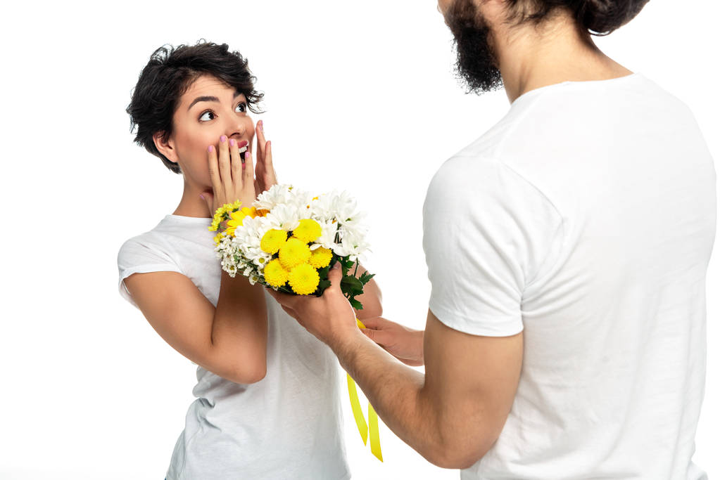 περικομμένη θέα του άντρα με γένια δίνοντας λουλούδια σε έκπληκτη Λατινική γυναίκα απομονώνεται σε λευκό  - Φωτογραφία, εικόνα