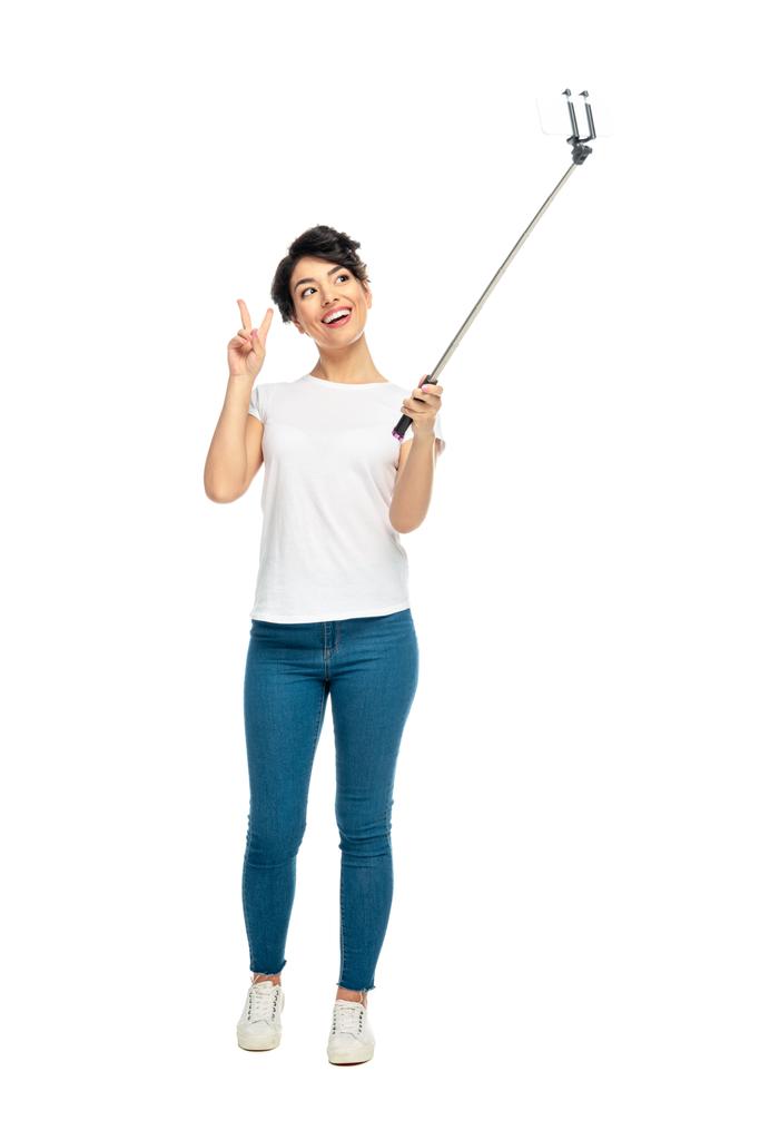 fröhliche lateinische Frau zeigt Friedenszeichen, während sie einen Selfie-Stick in der Hand hält und ein Selfie isoliert auf Weiß macht  - Foto, Bild