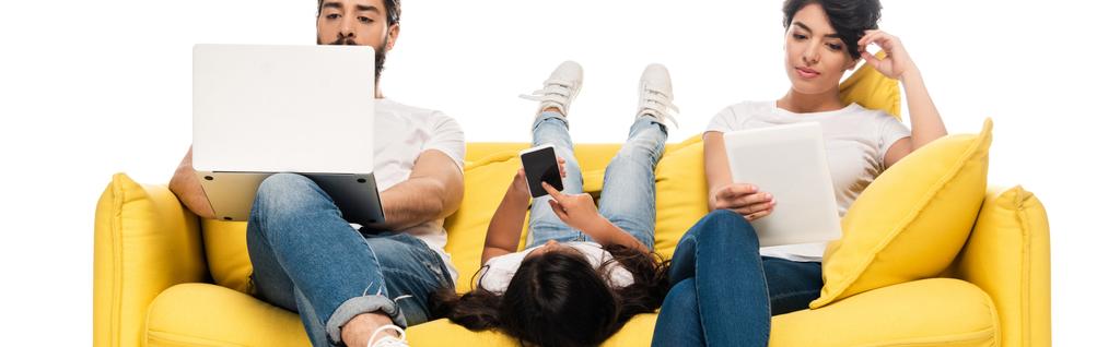 plan panoramique d'un enfant allongé sur un canapé et tenant un smartphone avec écran vide près de parents latins à l'aide de gadgets isolés sur du blanc
  - Photo, image