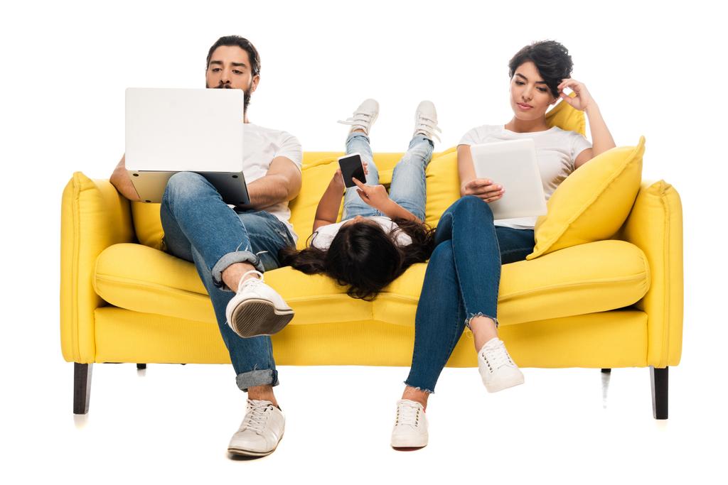 niño acostado en el sofá y sosteniendo el teléfono inteligente con pantalla en blanco cerca de los padres latinos utilizando gadgets aislados en blanco
  - Foto, Imagen