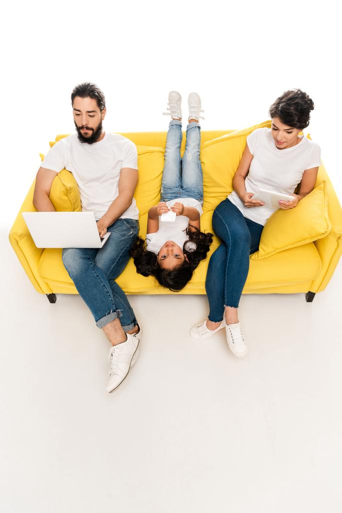 yleisnäkymä lapsi makaa sohvalla ja tilalla älypuhelin lähellä latino vanhemmat käyttävät vempaimia eristetty valkoinen
   - Valokuva, kuva