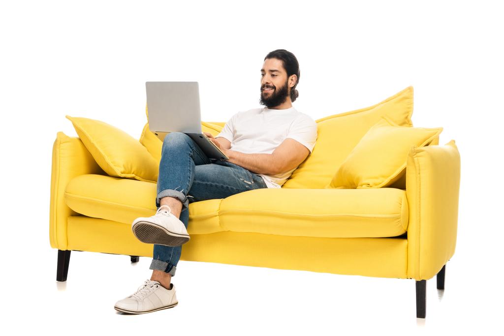 щасливий бородатий латинський чоловік у блакитних джинсах сидить у жовтому дивані і використовує ноутбук ізольований на білому
  - Фото, зображення