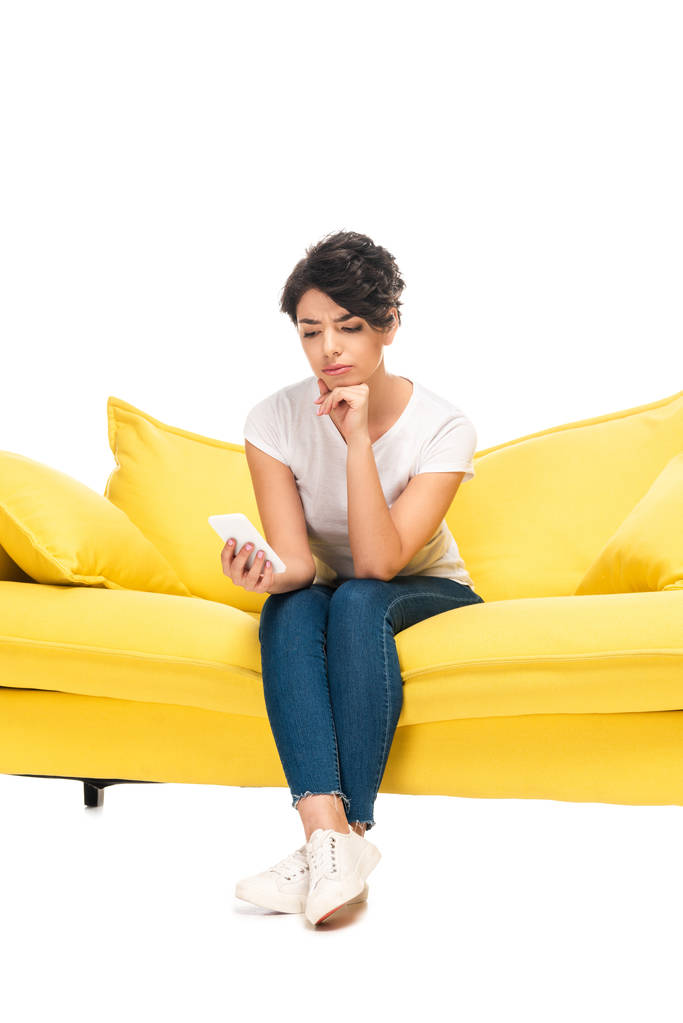 αναστατωμένη Λατινική γυναίκα που κοιτάζει το smartphone, ενώ κάθεται σε καναπέ απομονώνεται σε λευκό  - Φωτογραφία, εικόνα