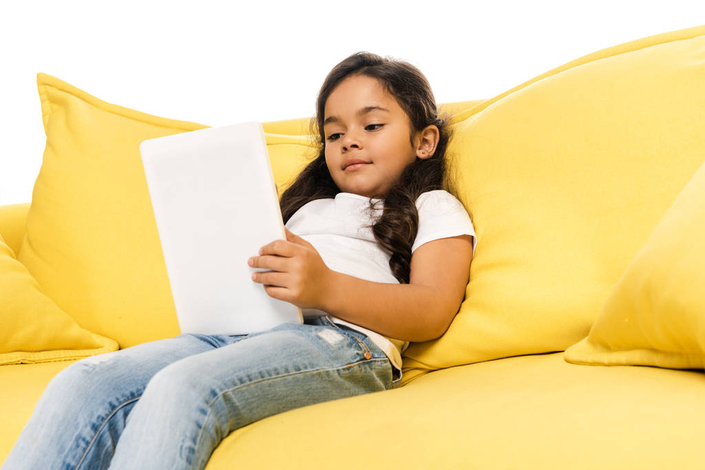 низький кут зору латинської дитини, використовуючи цифровий планшет, сидячи на дивані ізольовано на білому
  - Фото, зображення