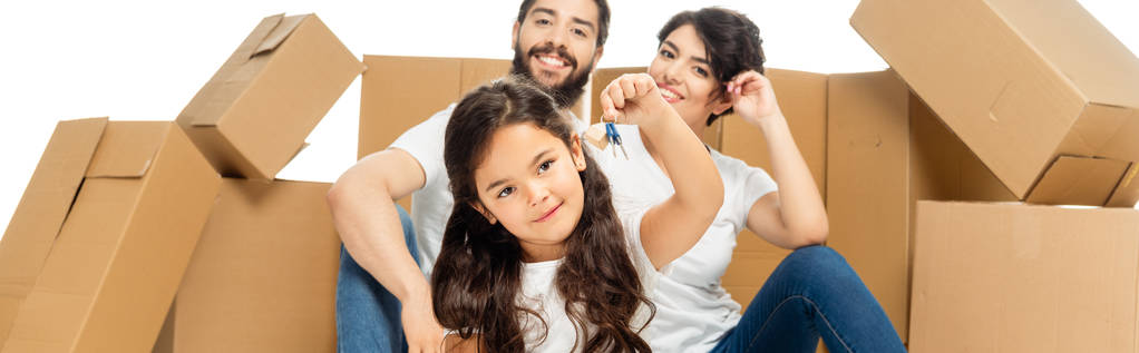 panoramic shot of of happy latin kid holding keys near parents and boxes isolated on white  - Photo, Image