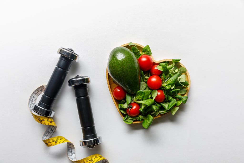 vista superior de las verduras en un tazón en forma de corazón, cinta métrica y pesas sobre fondo blanco
 - Foto, Imagen