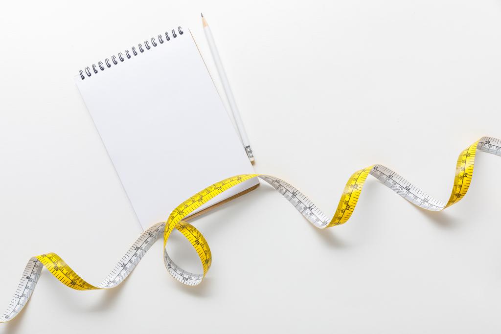 Maßband von oben, leeres Notizbuch mit Bleistift auf weißem Hintergrund  - Foto, Bild