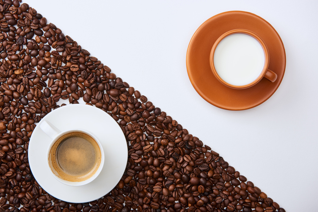top view of delicious coffee in cup among roasted beans near cup of milk on white background - Photo, Image