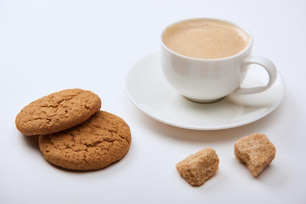 вкусный кофе с пеной в чашке на блюдце рядом с коричневым сахаром и печенье на белом фоне
 - Фото, изображение