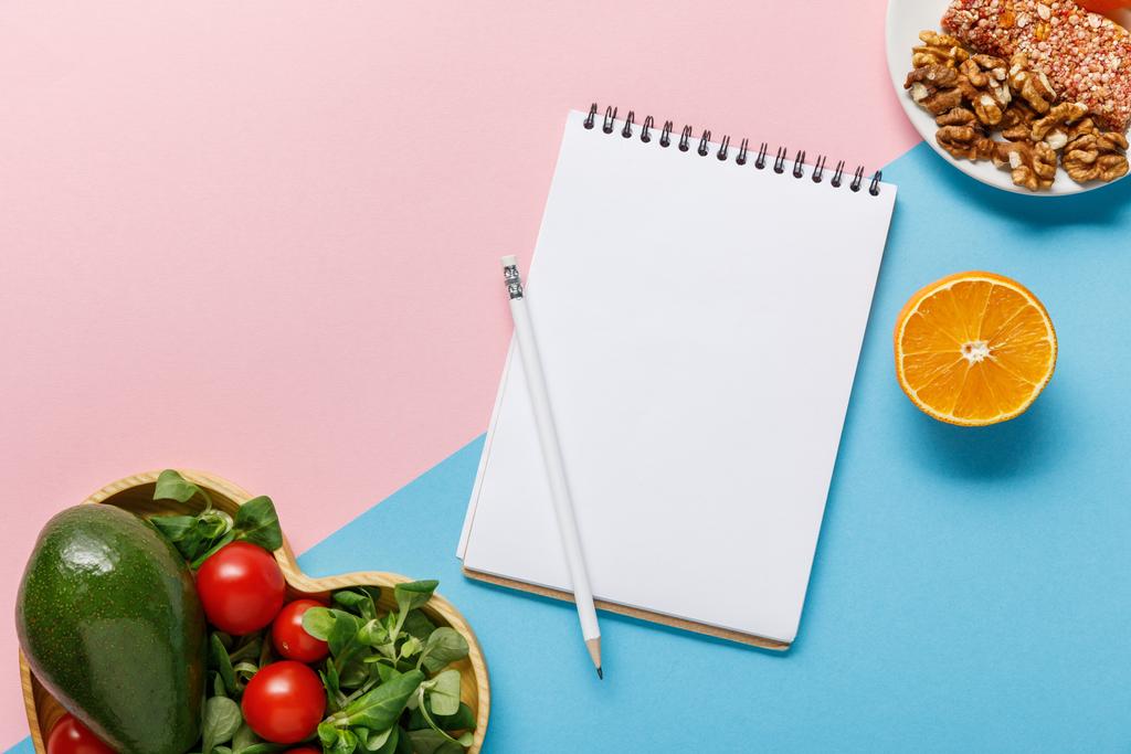 top view of empty notebook, vegetable salad, orange and nuts on pink and blue background - 写真・画像