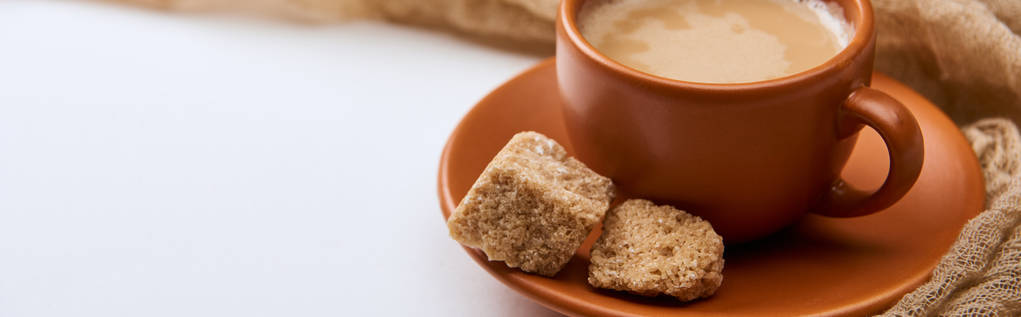 plano panorámico de sabroso café con espuma en taza en platillo cerca de azúcar morena sobre fondo blanco con tela
 - Foto, Imagen
