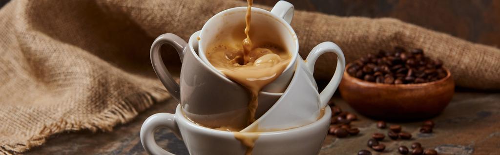 panoramische shot van stromend hete koffie uit kopjes op marmeren oppervlak in de buurt van zak en bonen - Foto, afbeelding