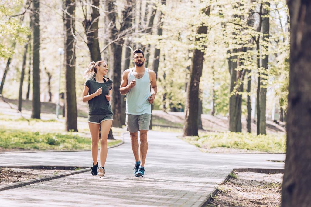 knappe man en mooie vrouw glimlachend tijdens het hardlopen in het groene zonnige Park  - Foto, afbeelding