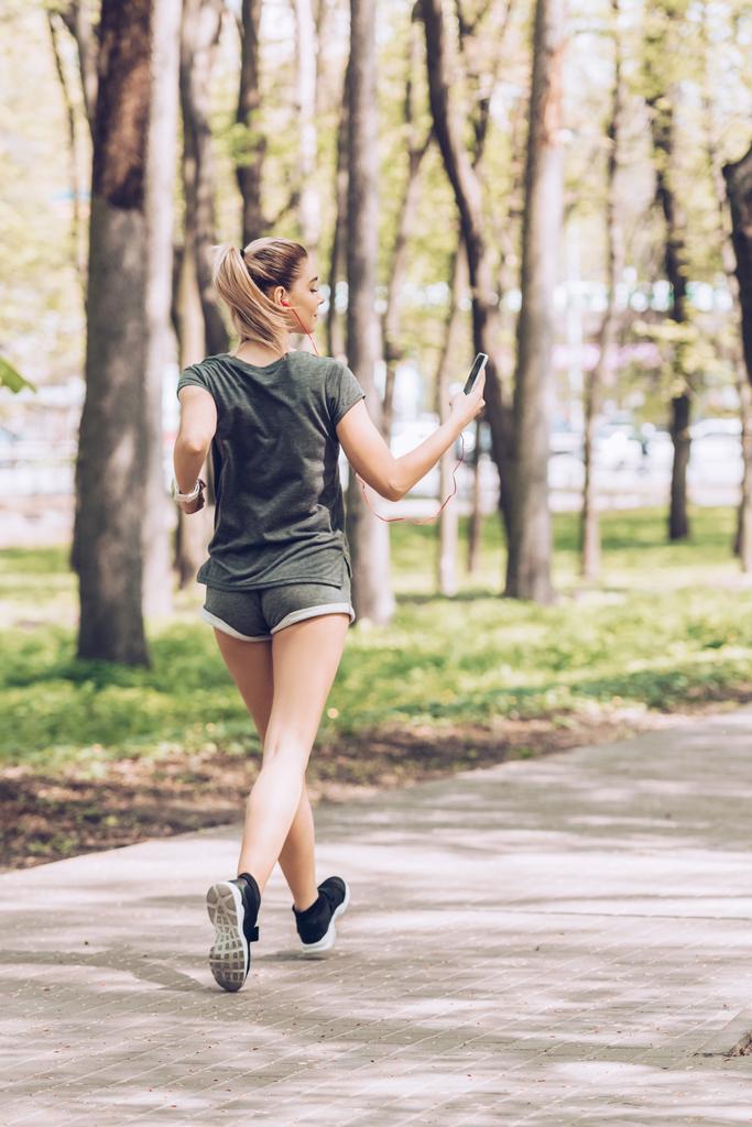 公園でジョギングをしながら、スマートフォンを持ち、イヤホンで音楽を聴いている若い女性のバックビュー - 写真・画像