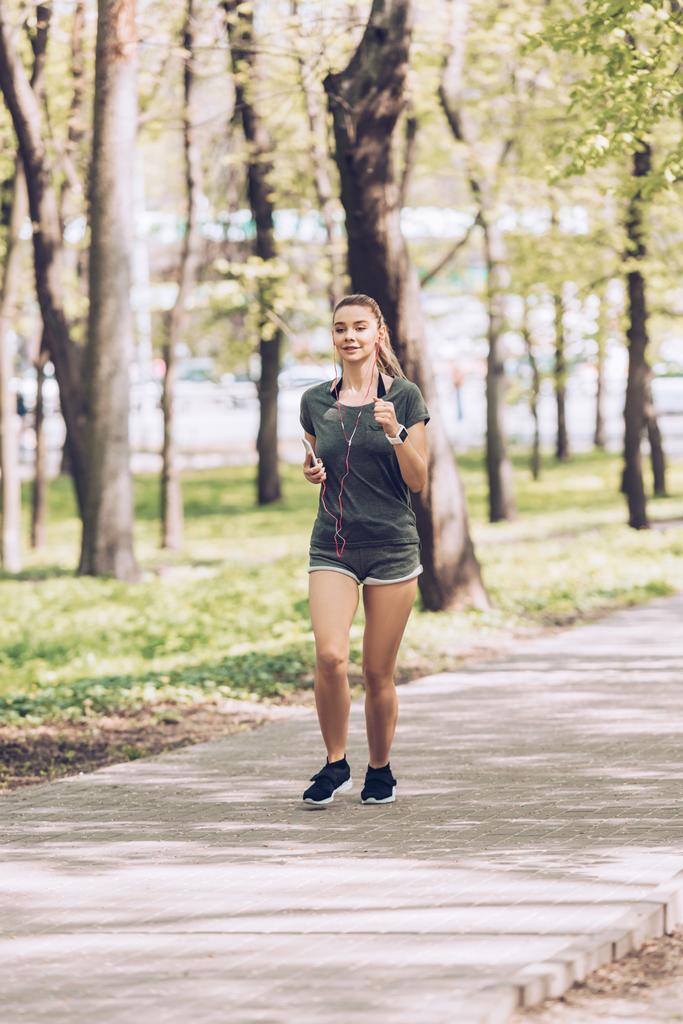 pretty young woman holding smartphone and listening music in earphones while running in park - Photo, Image