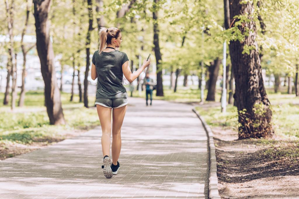 back view of young sportswoman jogging in park while holding smartphone and listening music in earphones - Photo, Image