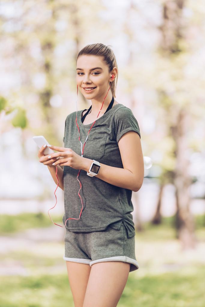 atrakcyjna kobieta w odzieży sportowej przy użyciu smartfona i słuchania muzyki w słuchawkach podczas uśmiechu w aparacie  - Zdjęcie, obraz