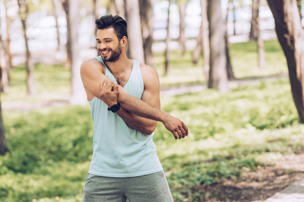 fröhlicher junger Sportler lächelt beim Stretching im sonnigen Park - Foto, Bild